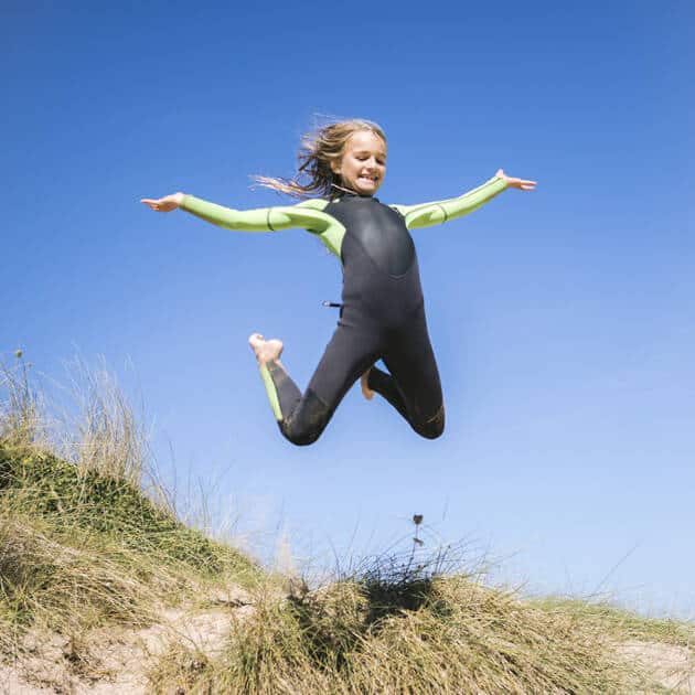 Wearing the C-Skins Junior Element 3/2mm Back Zip Wetsuit, a young person leaps joyfully with arms wide on a grassy sand dune beneath a clear blue sky.