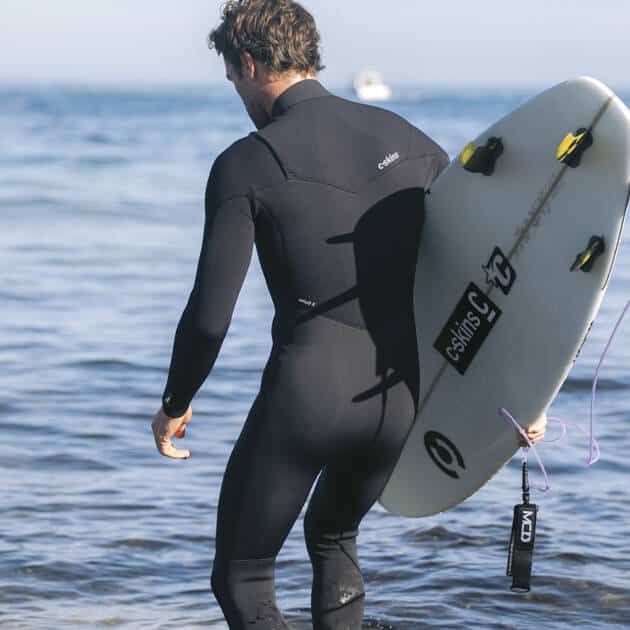 Wearing the C-Skins Men's ReWired 4/3mm Chest Zip Wetsuit, a person stands at the sea's edge holding a white surfboard, facing gentle waves breaking near their feet.