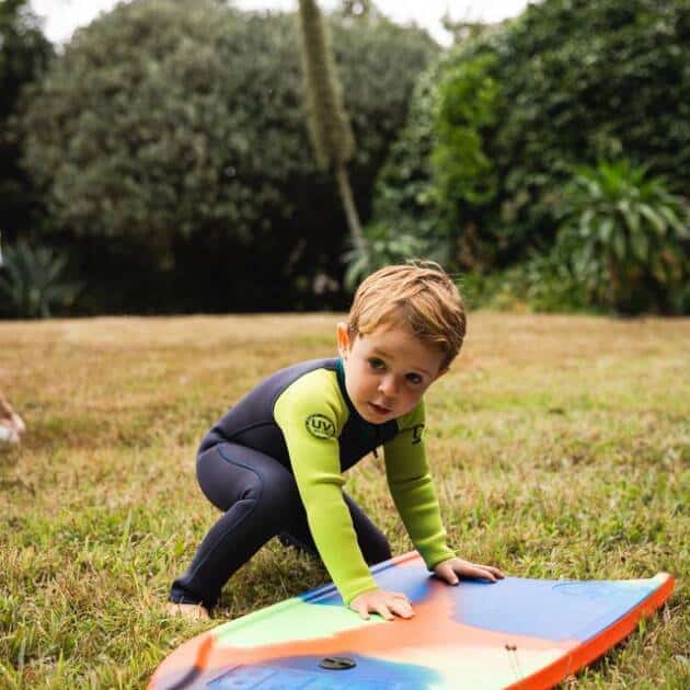 A young child wearing the C-Kid NuWave Baby Wetsuit kneels on grass and touches a colourful bodyboard with an orange edge, outdoors among trees and bushes.