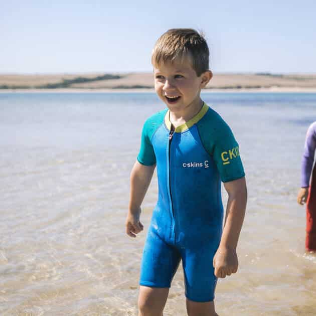A young boy wearing the C-Kid Baby Shorti Wetsuit smiles in shallow beach water, with another child partly visible behind him and sand dunes in the background.
