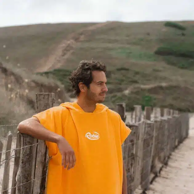 A person wearing a Robie Towel Robe stands by a wooden fence on a sandy beach, with grassy hills and a cloudy sky in the background.