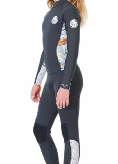 A smiling young girl with long wavy hair models the Ripcurl Junior Girls Dawn Patrol 5/3mm Back Zip Wetsuit, featuring black with white and floral accents, standing barefoot in profile against a white background.