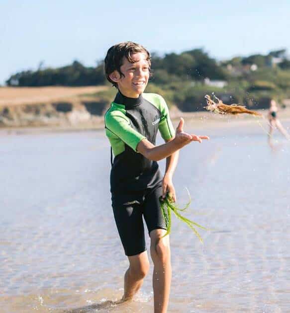 A smiling boy wearing the C-Skins Junior Element 3/2mm Shortie Wetsuit stands in shallow beach water, tossing seaweed with one hand under a clear sky with distant trees and hills in the background.