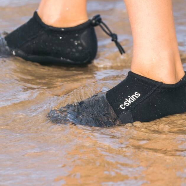 A person wearing C-Skins Legend 3mm Round Toe Slippers stands in shallow beach water, the product name clearly visible on the side of their black water shoes.