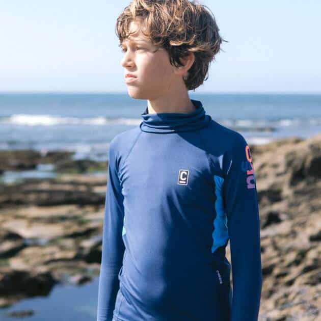 A young boy wearing the C-Skins NuWave Rash X LS Vest stands on a rocky shoreline, gazing into the distance with the sea and sky in the background.