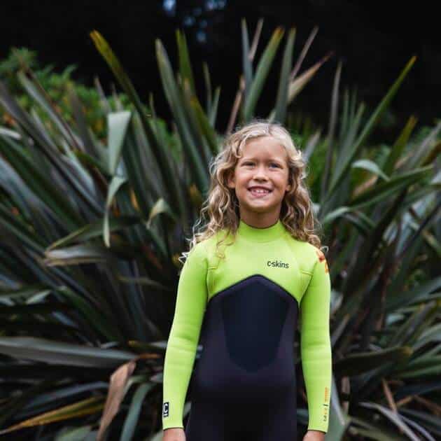 A smiling child with long, curly blond hair wears the C-Skins Junior NuWave Legend 4/3mm Back Zip Wetsuit whilst standing outdoors in front of tall, spiky green plants.
