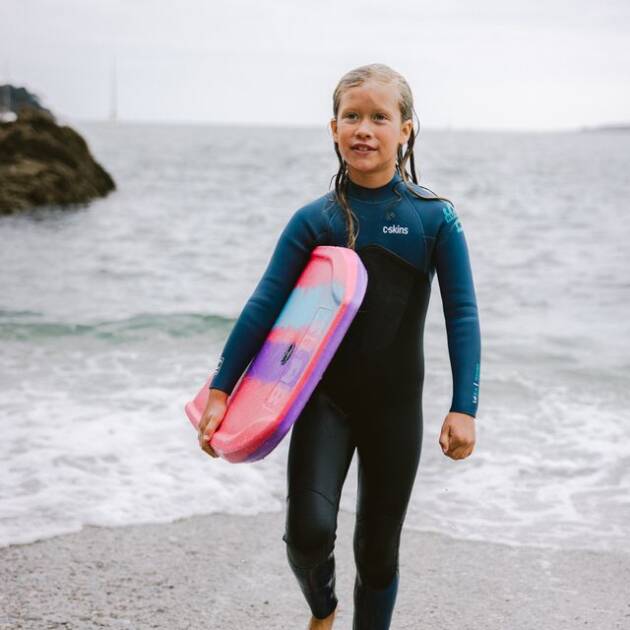 A young child wearing the C-Skins Junior NuWave Legend 5/4/3mm Back Zip Wetsuit walks barefoot on the beach, carrying a pink and purple bodyboard, with waves and a rocky shoreline in the background.