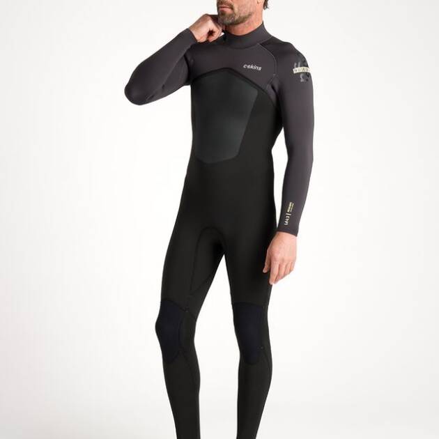 A man with short hair and a beard stands barefoot on a white background, wearing the C-Skins NuWave Legend 4/3mm Back Zip Wetsuit, which is black with long sleeves and a high collar.
