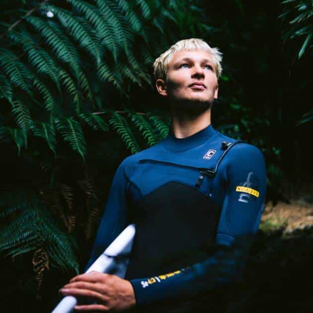 Wearing the C-Skins NuWave Session 4/3mm Chest Zip Wetsuit, a person holding a surfboard stands among green ferns, gazing upwards with a thoughtful look.