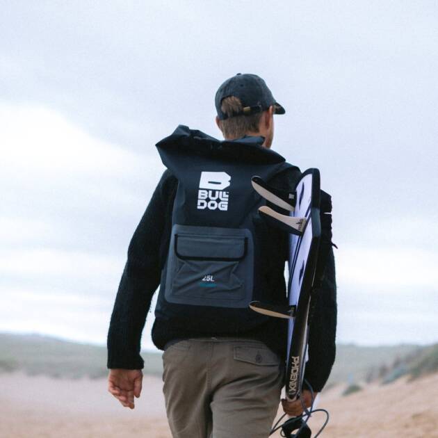 A person in a black cap and jumper carries a Bulldog Dry Rucksack and a hydrofoil board while walking outdoors on sandy terrain under a cloudy sky.