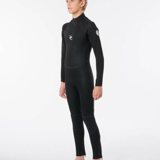 A young person with short blonde hair stands barefoot on a white background, wearing the Ripcurl Junior Freelite 5/3mm Back Zip Wetsuit, a black full-body wetsuit with long sleeves and small white logos on the chest and upper arm.