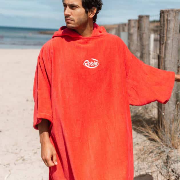 Wearing a loose, bright red Robie Long Sleeve Towel Robe with "Robie" on the chest, a person stands on a sandy beach near a wooden fence, gazing into the distance with the sea and other beachgoers in the background.