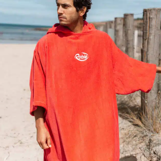 Wearing a loose, bright red Robie Long Sleeve Towel Robe with "Robie" on the chest, a person stands on a sandy beach near a wooden fence, gazing into the distance with the sea and other beachgoers in the background.