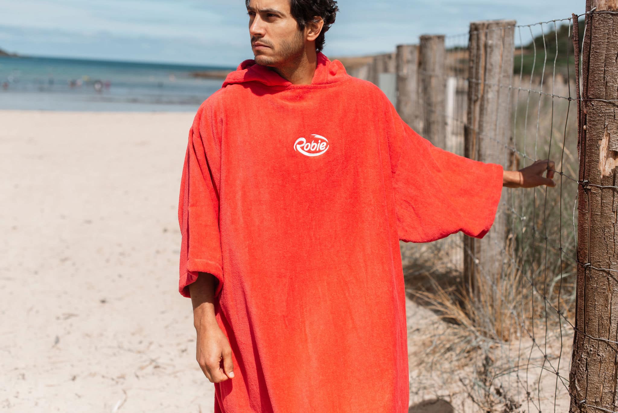 Wearing a loose, bright red Robie Long Sleeve Towel Robe with "Robie" on the chest, a person stands on a sandy beach near a wooden fence, gazing into the distance with the sea and other beachgoers in the background.