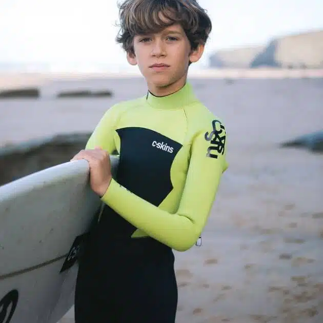 A young boy wears the C-Skins Junior Legend 5/4/3mm Back Zip Wetsuit on a sandy beach, holding a surfboard. The sea and rocky cliffs can be seen in the background under a partly cloudy sky.