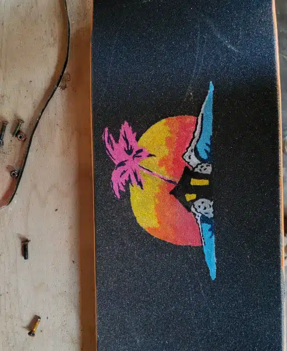 A skateboard deck with black grip tape features a colourful sunset, palm tree, and ocean design. Nearby are skateboard parts, tools, a pink wheel, and a person’s tan trousers and shoes visible at the edge of the image.