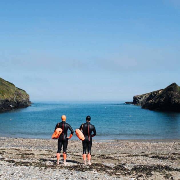 Two people in wetsuits and orange swim caps stand on a rocky beach, each holding a C-Skins Swim Research Buoy Bag 28L, facing a calm blue sea bordered by cliffs under a clear sky.