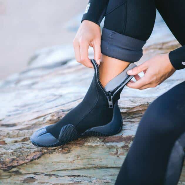 A person in a wetsuit sits on a rock, zipping up C-Skins Adult Legend 5mm Zipped Round Toe Boots over their ankle as they get ready for water activities.