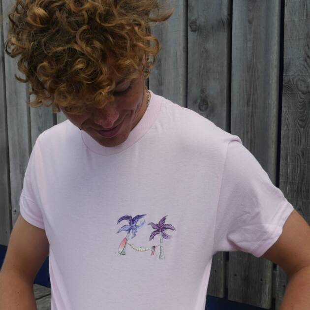A person with curly hair looks down while wearing the Shack Brand Palm Tree Adults' T-Shirt in light pink, featuring a small design of two palm trees and a hammock. Wooden boards form the background.