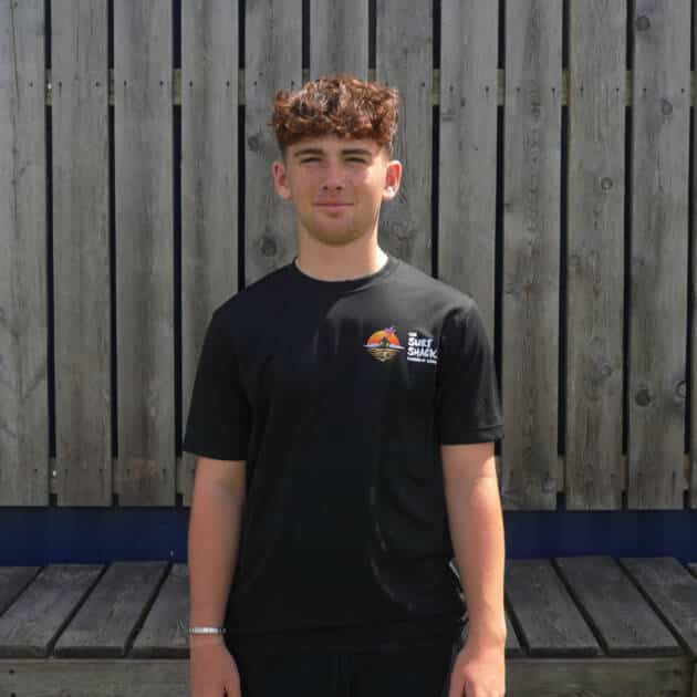 A young person with curly brown hair stands in front of a wooden fence, wearing The Surf Shack Sports T-Shirt featuring a colourful logo on the left chest, designed for breathability and comfort.