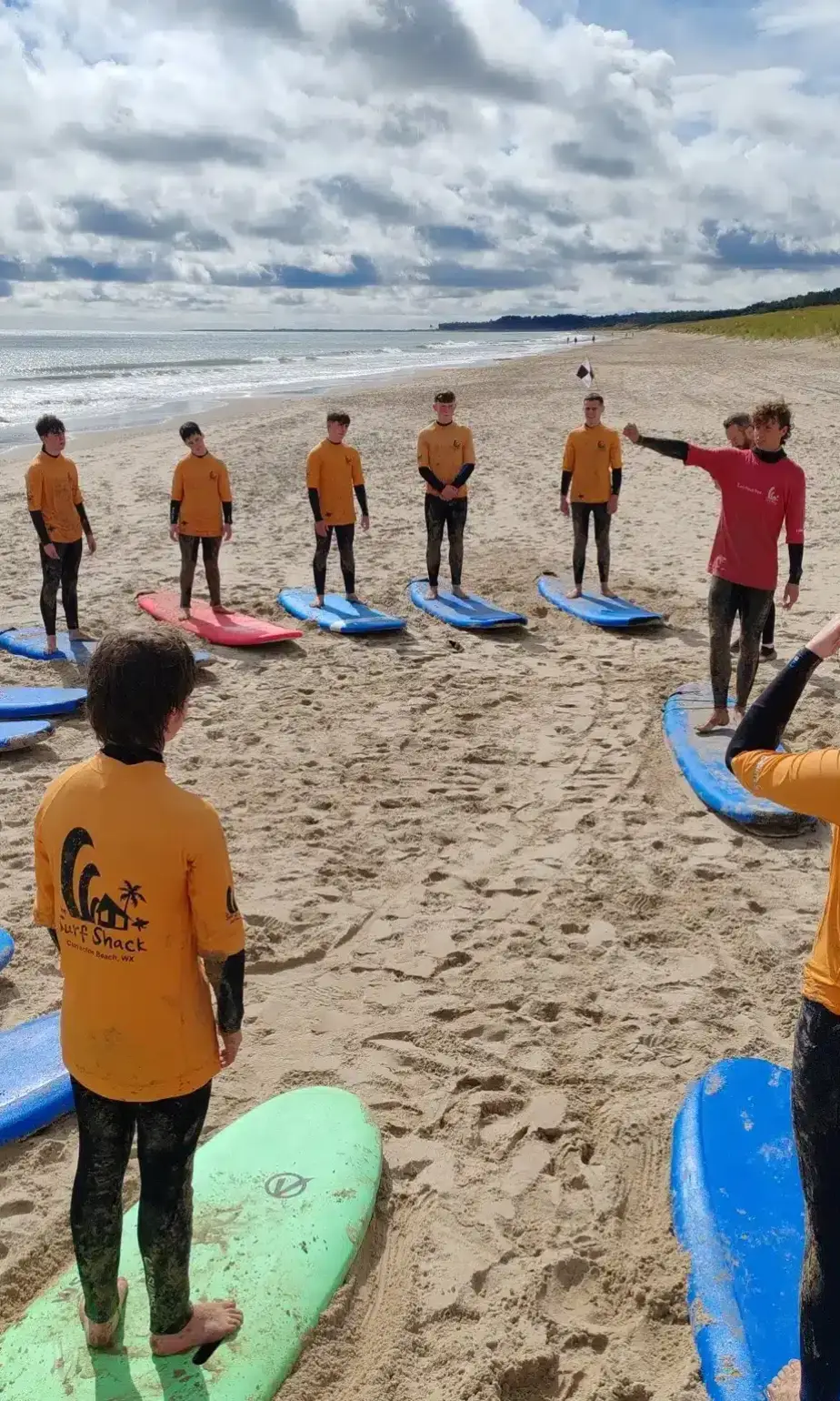 A group of children in yellow "Surf Shack" shirts stand on a sandy beach in a semicircle with surfboards, listening to two instructors giving instructions under a cloudy sky.