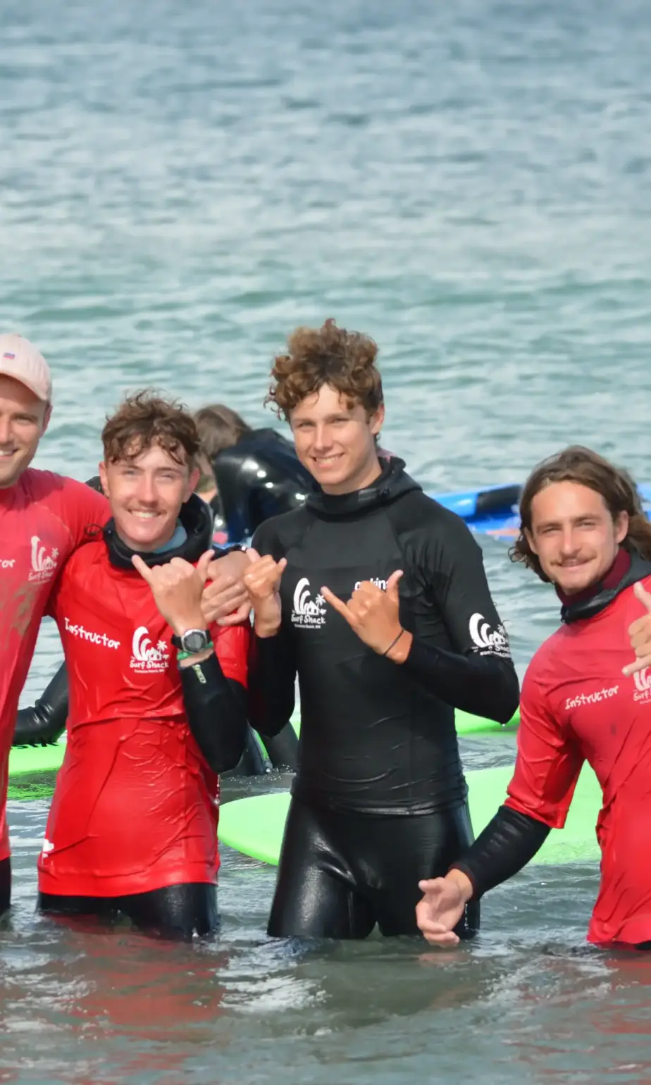 Four people in wetsuits and red instructor shirts stand in shallow water at the beach, smiling and making hand gestures, with surfboards and the sea visible in the background.